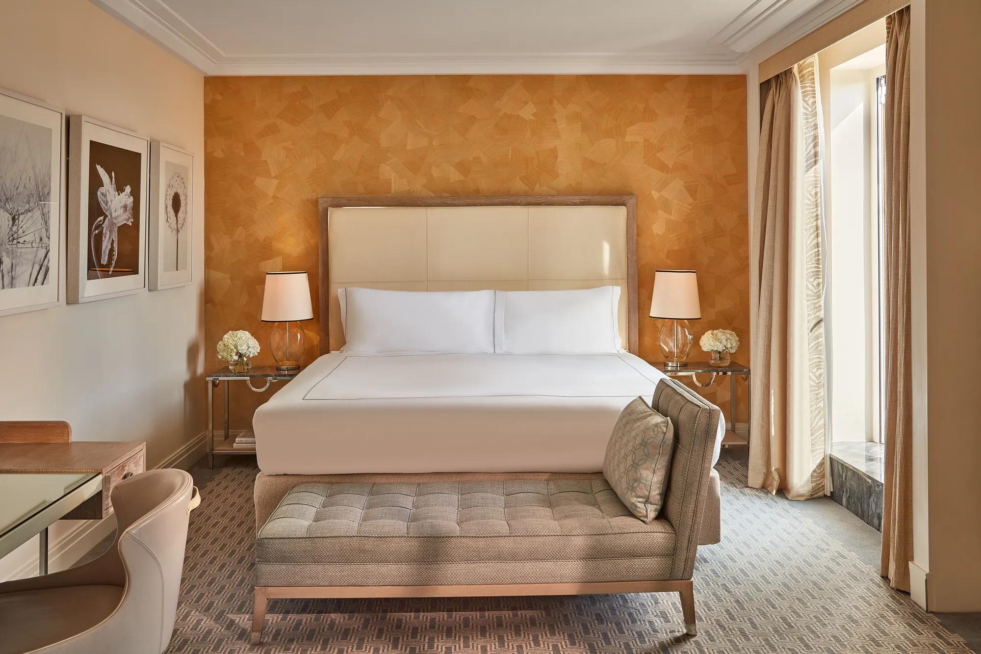 Hotel bedroom with cream headboard, patterned orange accent wall, and bench at foot of bed.