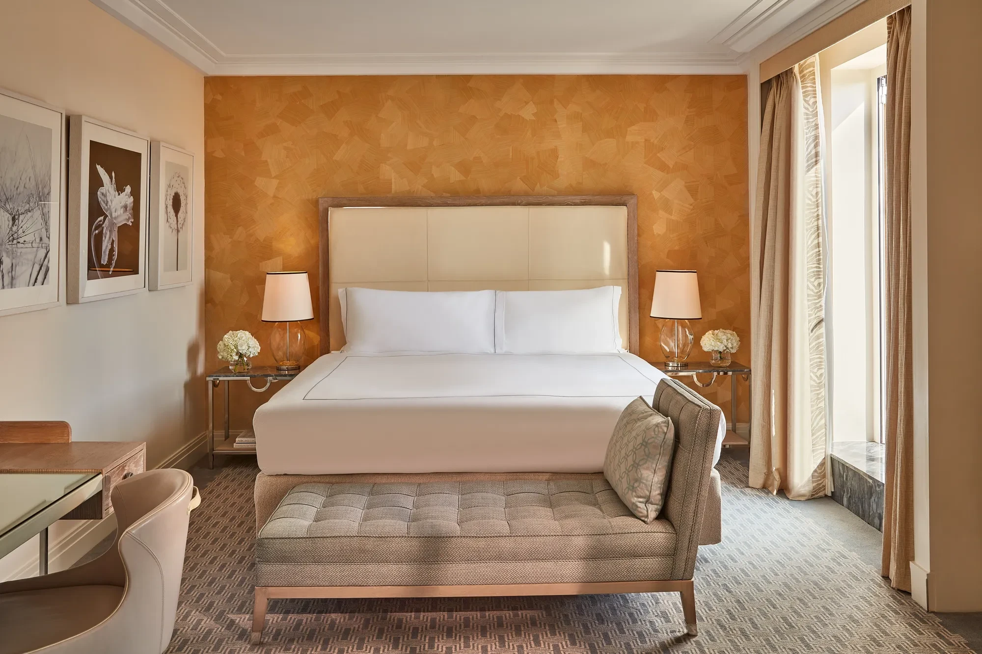 Hotel bedroom with cream headboard, patterned orange accent wall, and bench at foot of bed.