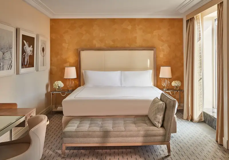 Hotel bedroom with cream headboard, patterned orange accent wall, and bench at foot of bed.