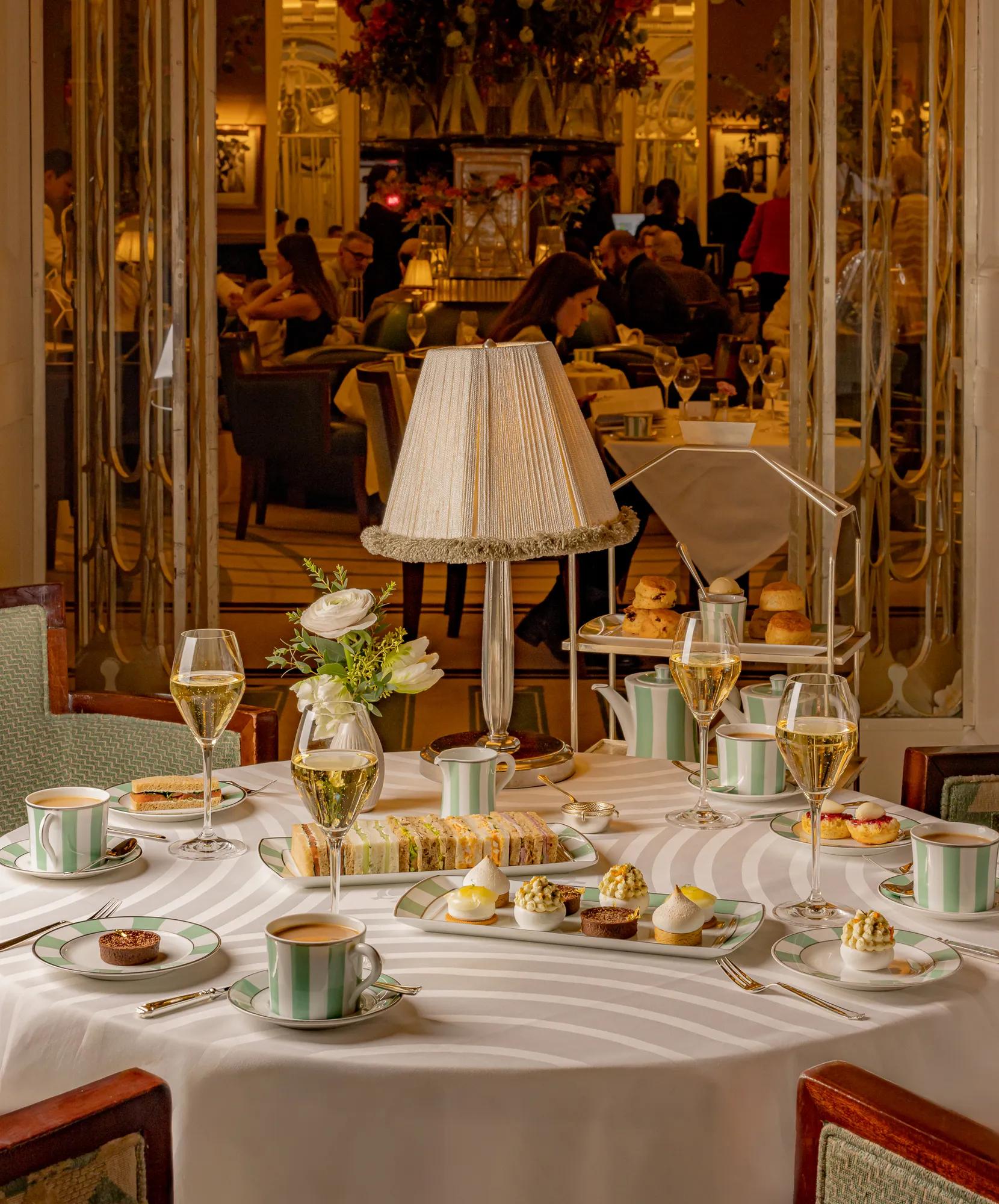 Elegant afternoon tea table set with striped china, delicate pastries, finger sandwiches, and champagne glasses arranged around a central table lamp with a fringed shade. The background shows a lively tea room with guests enjoying service in a refined, classic setting.