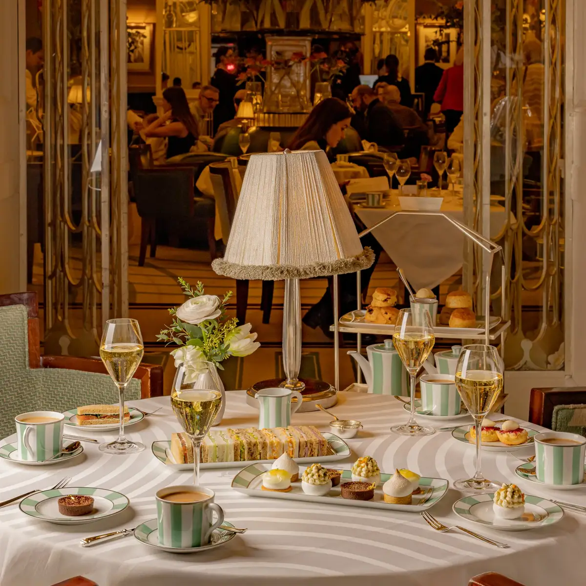 Elegant afternoon tea table set with striped china, delicate pastries, finger sandwiches, and champagne glasses arranged around a central table lamp with a fringed shade. The background shows a lively tea room with guests enjoying service in a refined, classic setting.