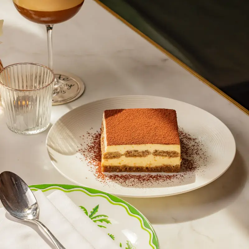 Slice of tiramisu dusted with cocoa on white plate beside an espresso martini on a marble café table.
