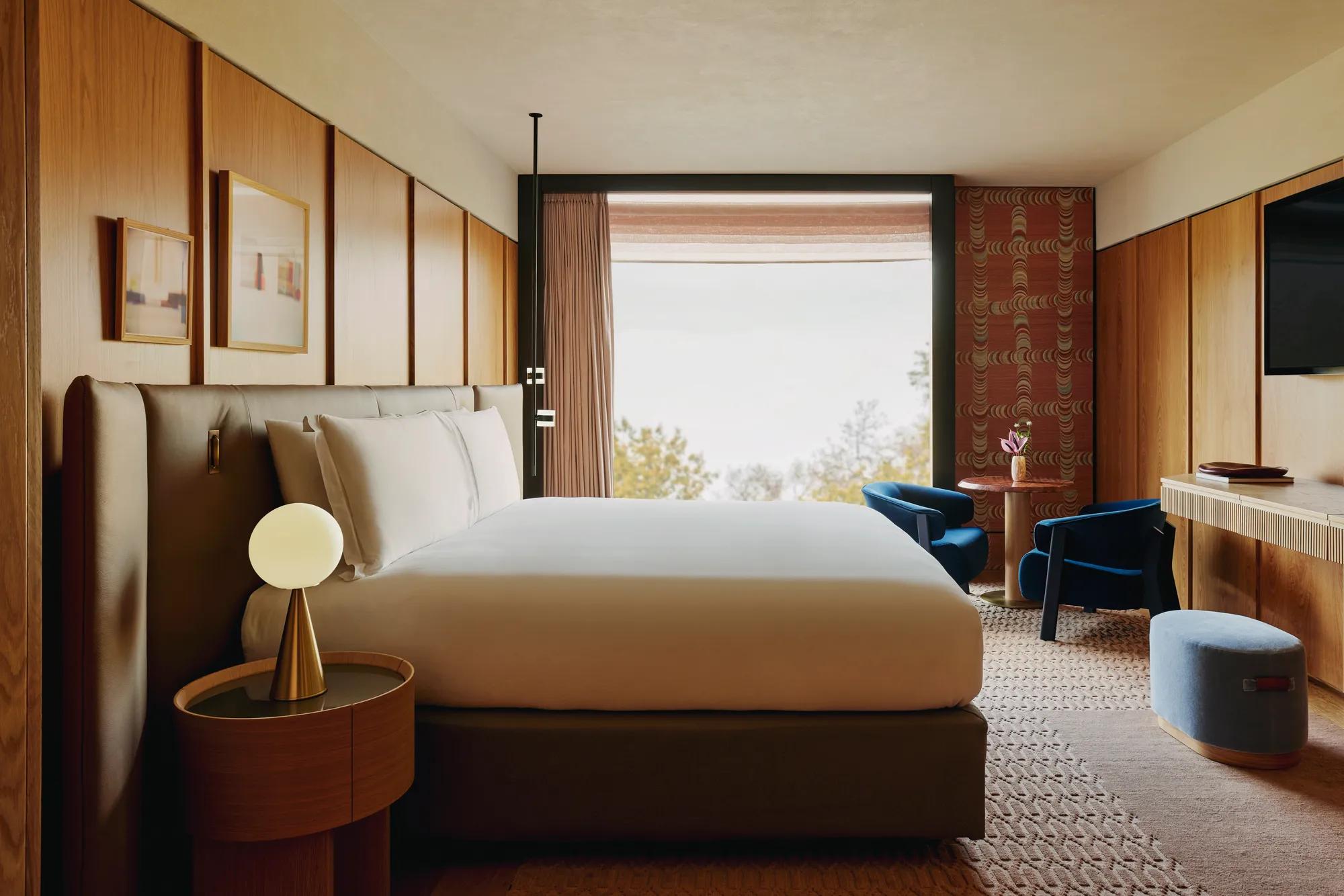 Bedroom with king-size bed, warm wood panelling, seating area, and large window overlooking treetops.