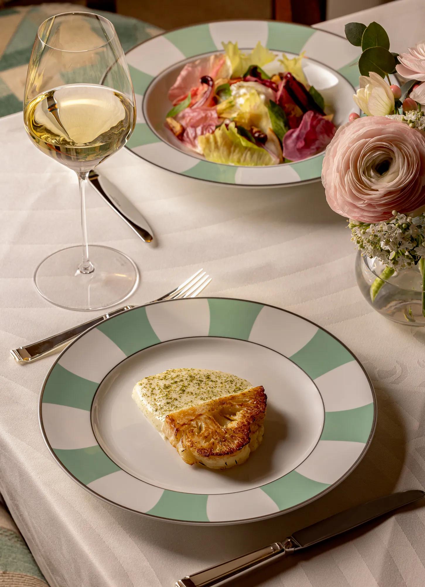 Elegant table setting with a plated dish of seared fish on a white sauce, served on green-and-white patterned china. A glass of white wine, a colorful salad, and a vase of pale pink flowers complete the refined dining scene.