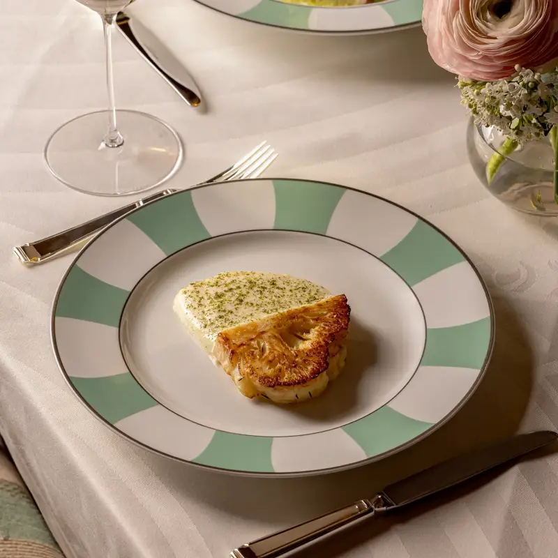 Elegant table setting with a plated dish of seared fish on a white sauce, served on green-and-white patterned china. A glass of white wine, a colorful salad, and a vase of pale pink flowers complete the refined dining scene.