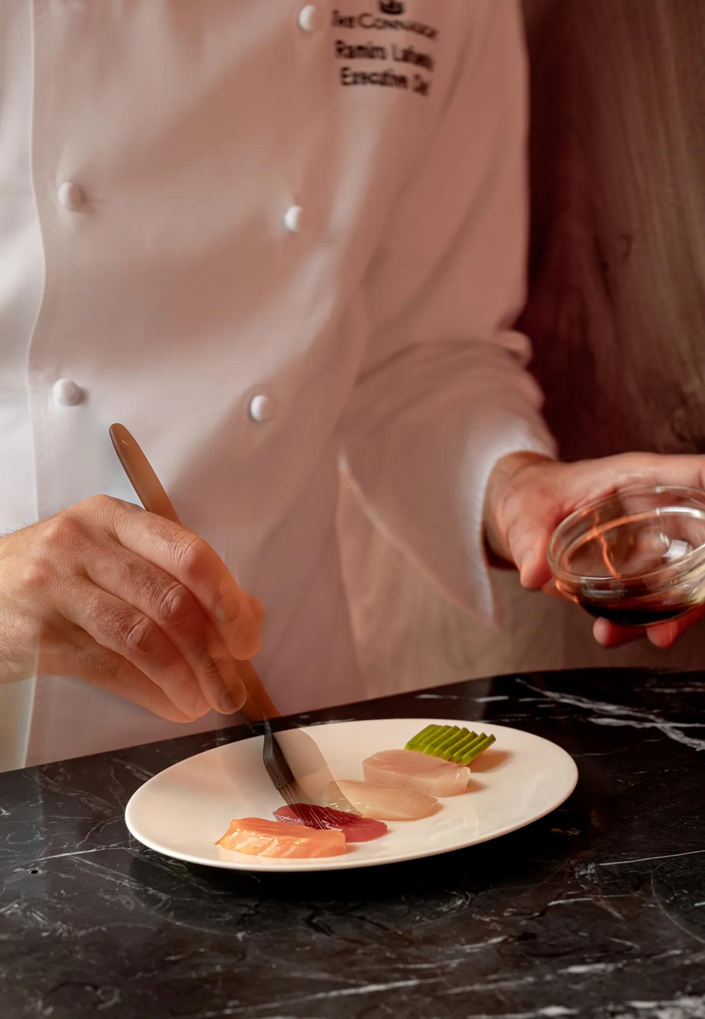 Chef carefully plating slices of sashimi and garnish on a white plate at a marble counter.