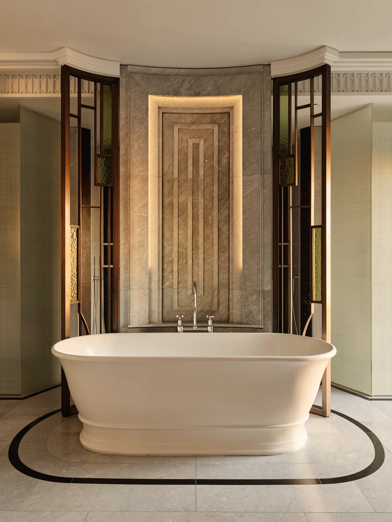 Luxurious bathroom featuring a freestanding oval bathtub set against a marble accent wall with geometric detailing and warm backlighting, framed by decorative glass and bronze panels.