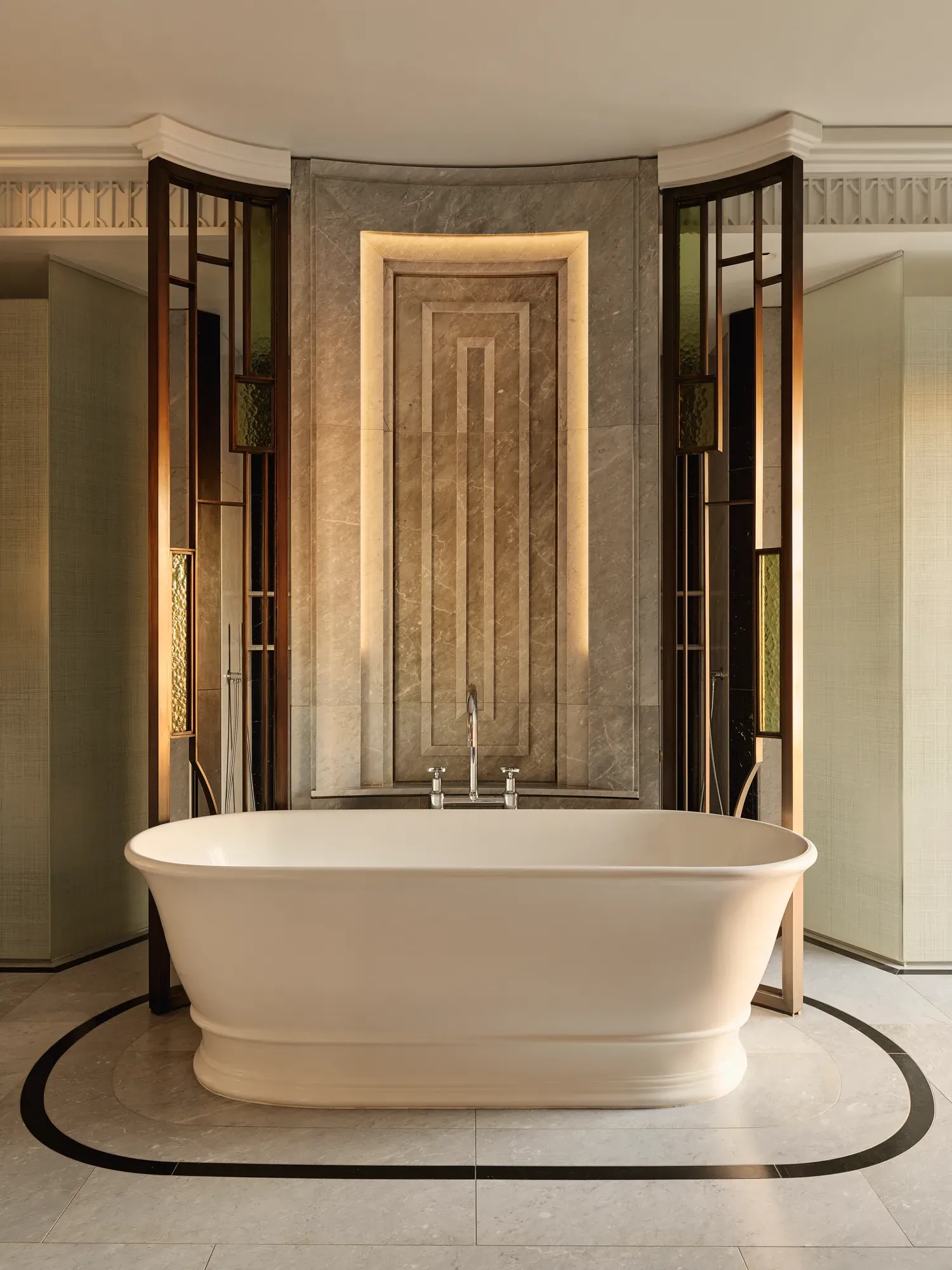 Luxurious bathroom featuring a freestanding oval bathtub set against a marble accent wall with geometric detailing and warm backlighting, framed by decorative glass and bronze panels.