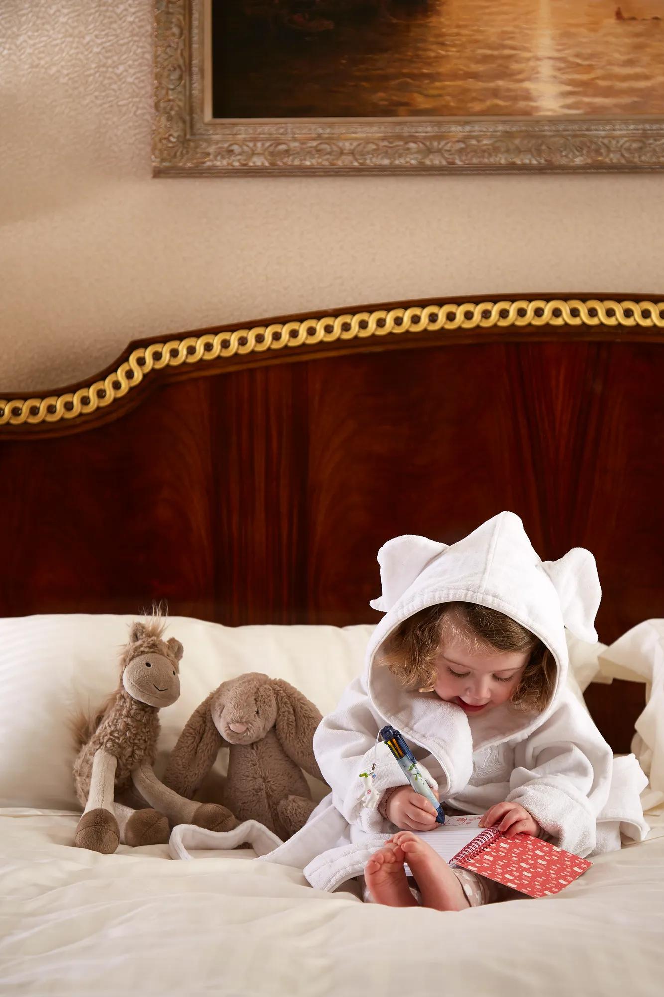 Child in white robe sitting on bed, drawing on card, with soft toys beside them against wooden headboard and framed artwork above.