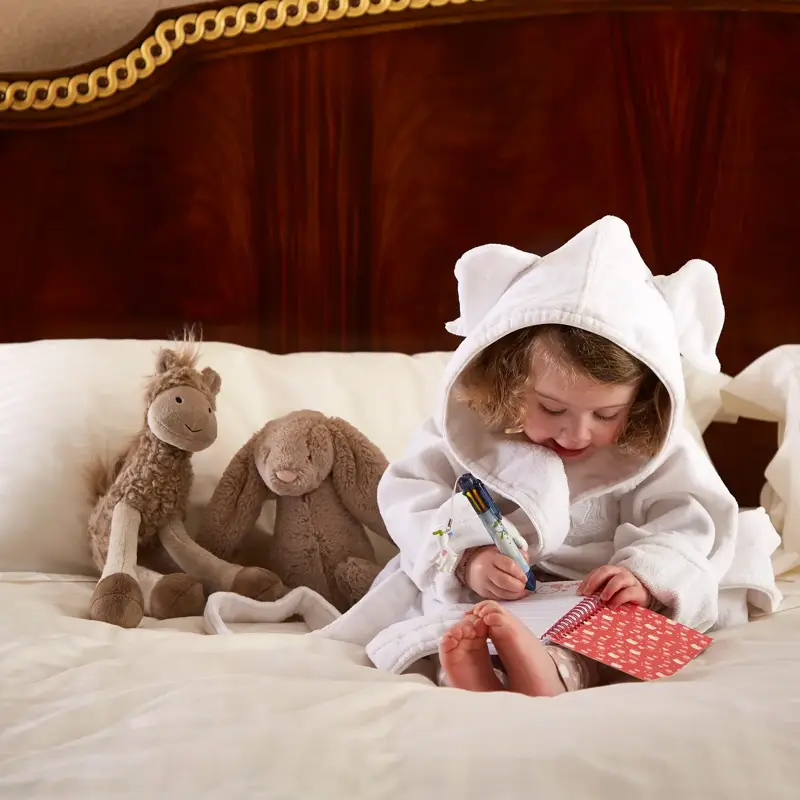 Child in white robe sitting on bed, drawing on card, with soft toys beside them against wooden headboard and framed artwork above.