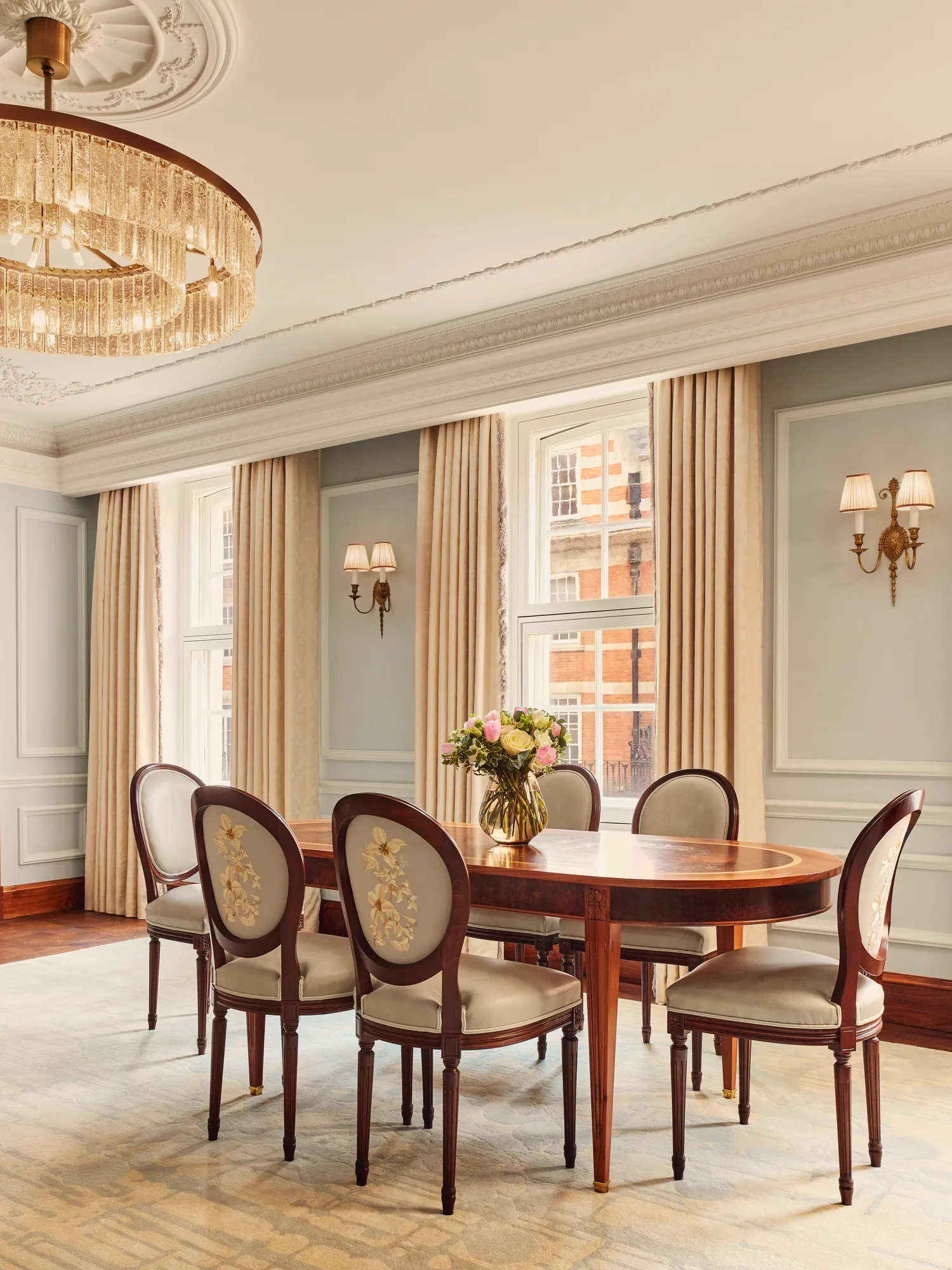 Refined dining area with an oval wooden table and six embroidered chairs, illuminated by a crystal chandelier and natural light from large windows with cream curtains.