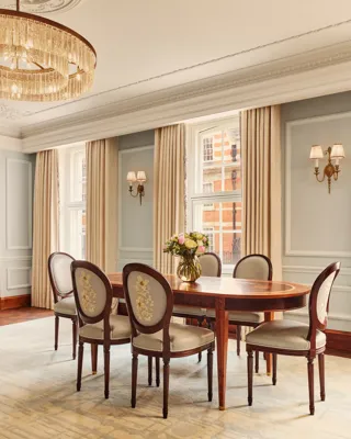 Refined dining area with an oval wooden table and six embroidered chairs, illuminated by a crystal chandelier and natural light from large windows with cream curtains.