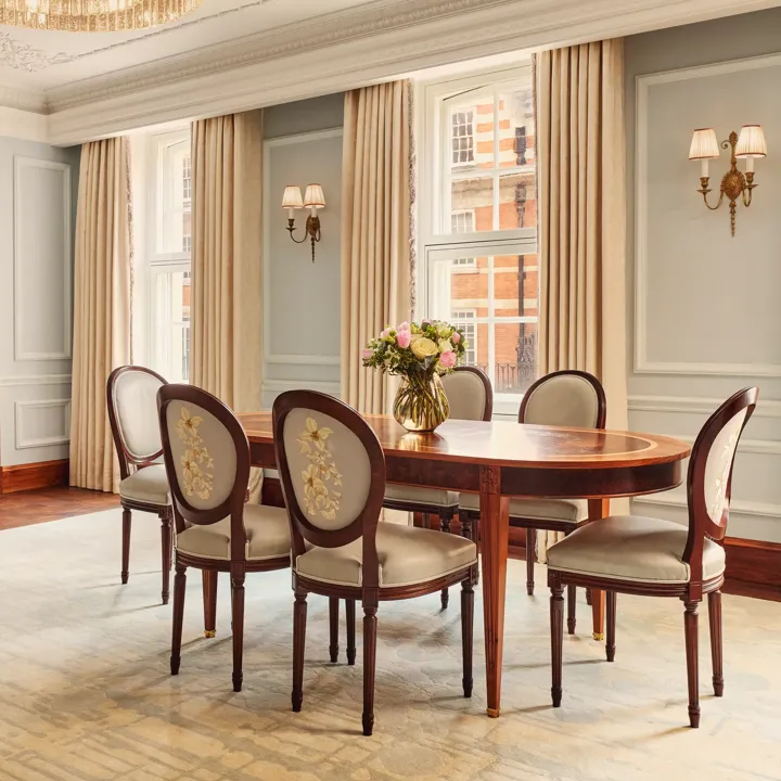 Refined dining area with an oval wooden table and six embroidered chairs, illuminated by a crystal chandelier and natural light from large windows with cream curtains.