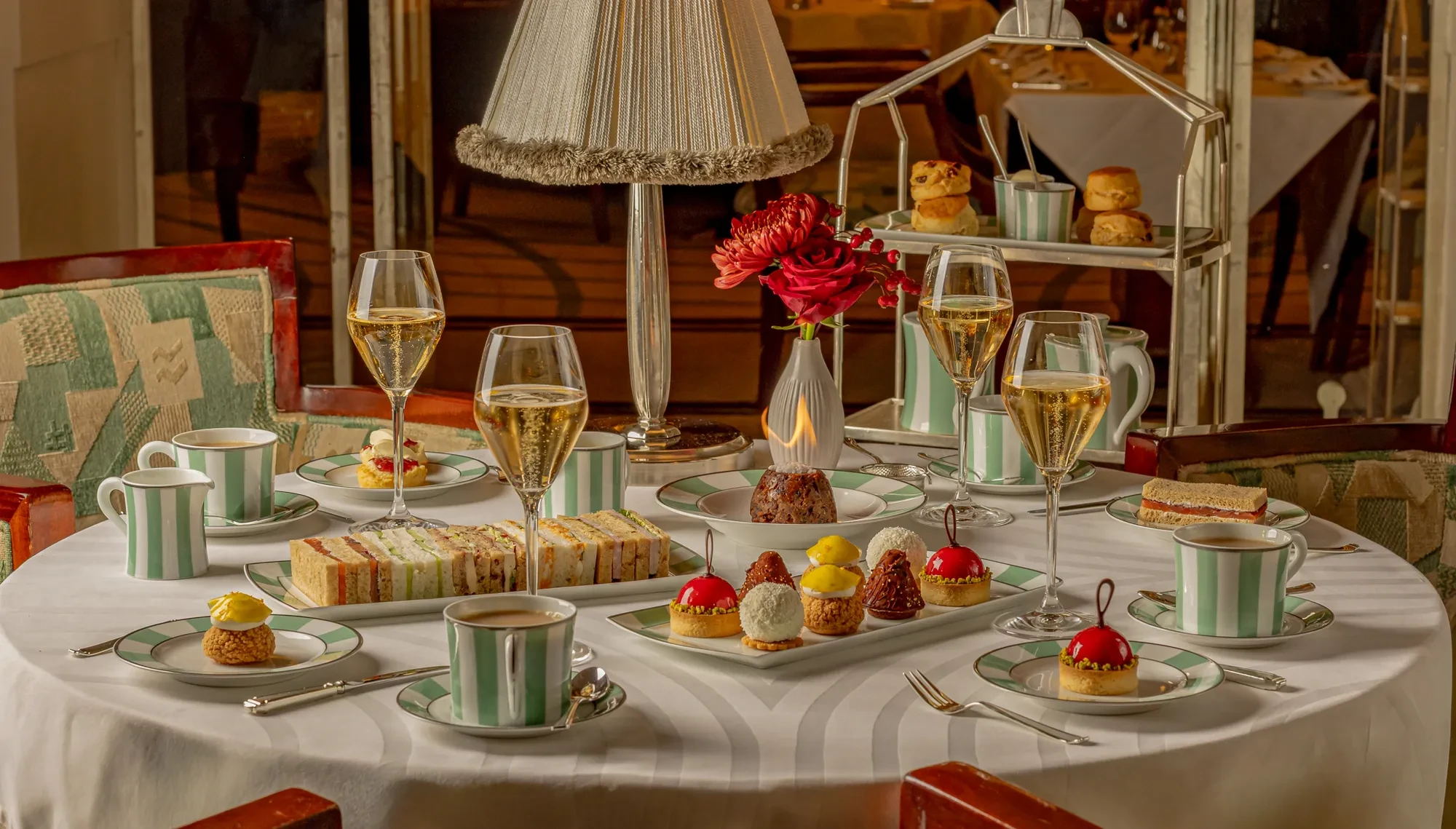 Elegant afternoon tea table with champagne flutes, finger sandwiches, pastries, striped china cups and a floral centrepiece.