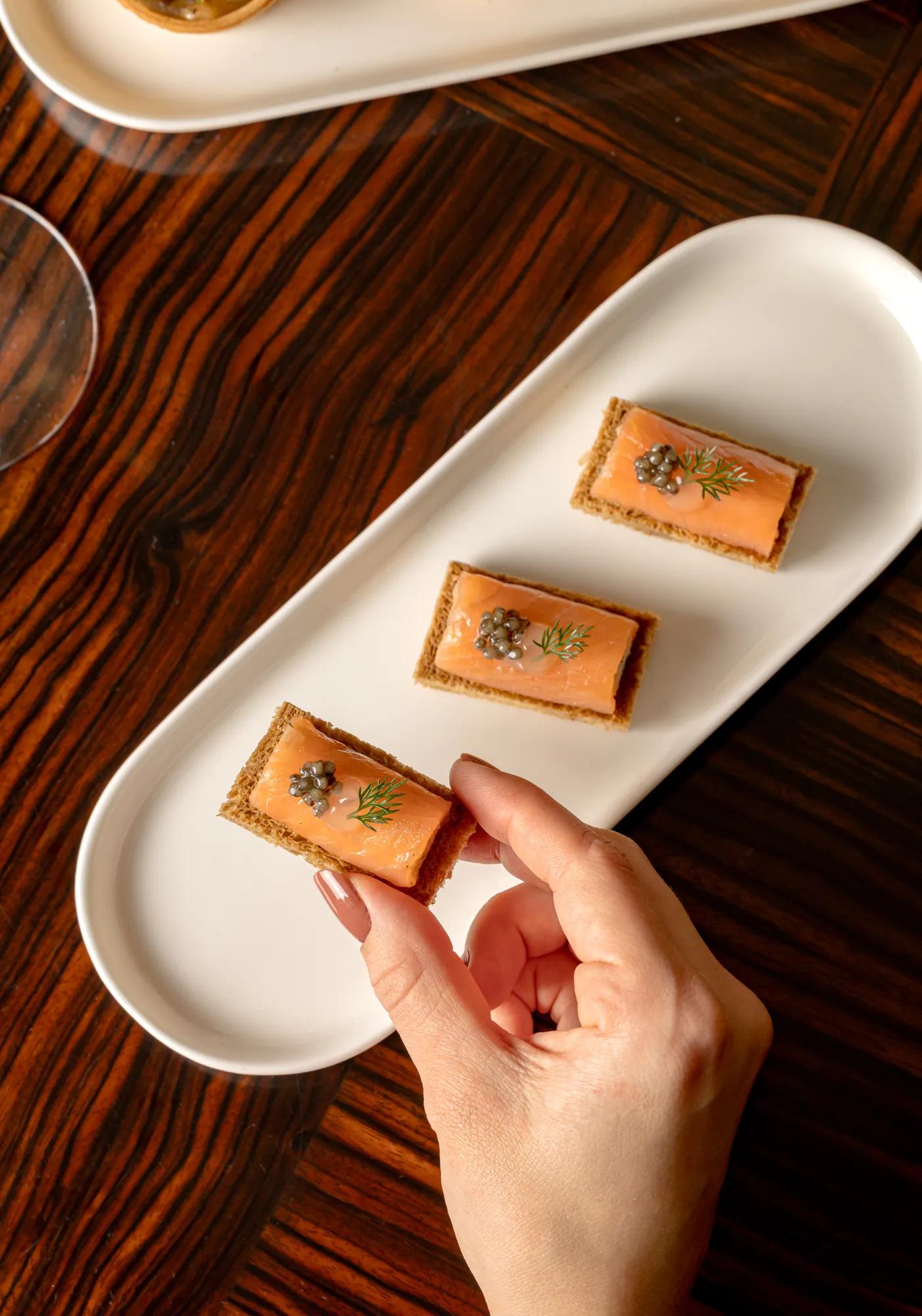 Hand picking smoked salmon canapé topped with caviar and dill from white serving platter on wooden table.