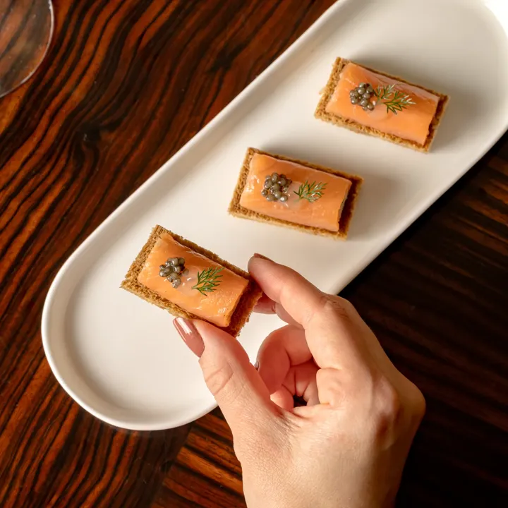 Hand picking smoked salmon canapé topped with caviar and dill from white serving platter on wooden table.