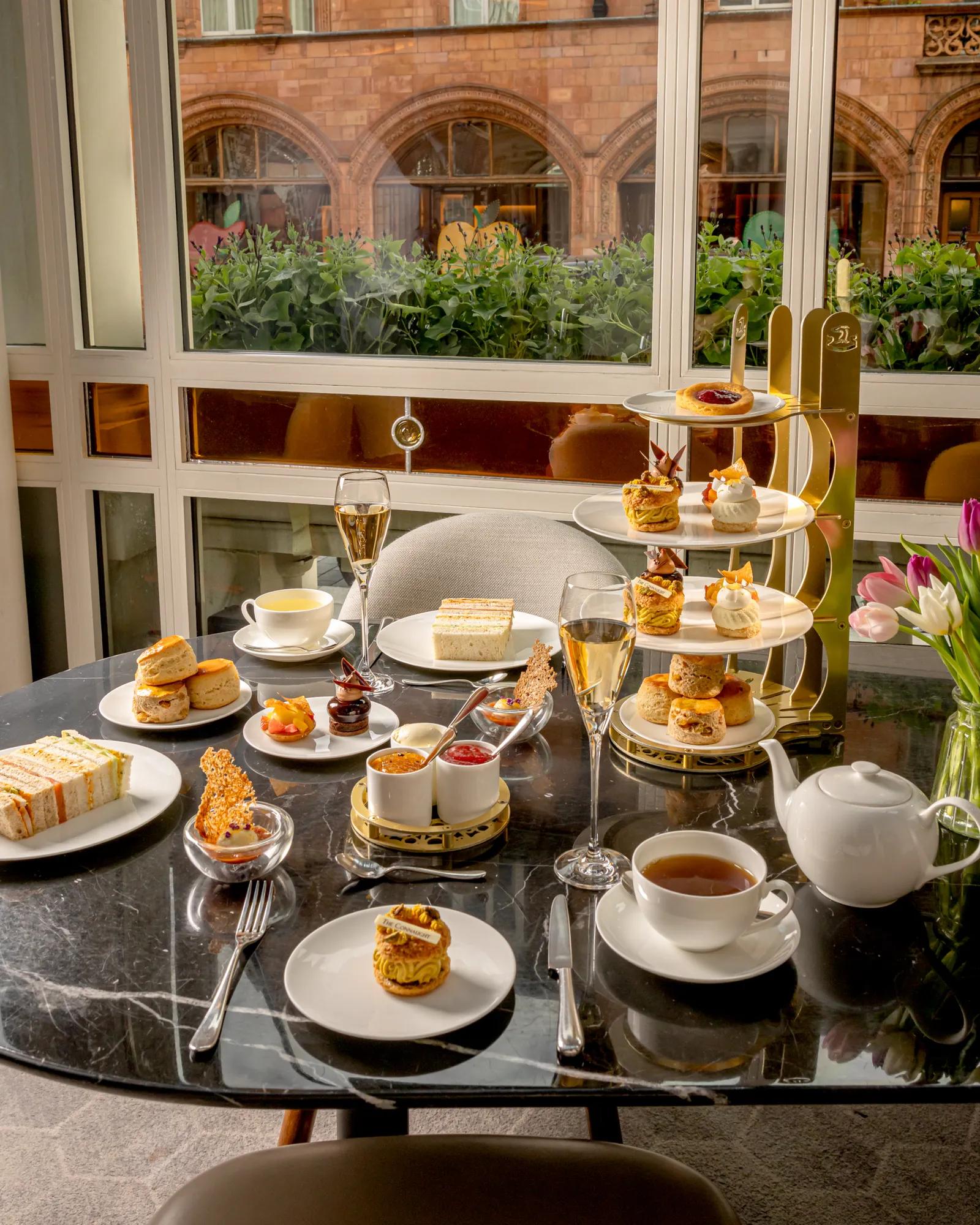 Afternoon tea spread with pastries, sandwiches, scones, tea, and champagne on a marble table by a window.