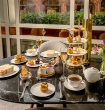 Afternoon tea spread with pastries, sandwiches, scones, tea, and champagne on a marble table by a window.