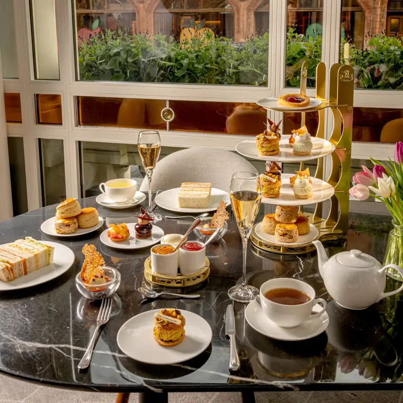 Afternoon tea spread with pastries, sandwiches, scones, tea, and champagne on a marble table by a window.