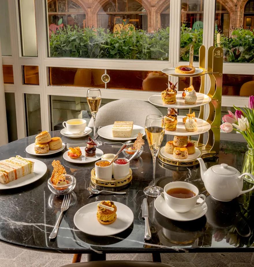 Afternoon tea spread with pastries, sandwiches, scones, tea, and champagne on a marble table by a window.