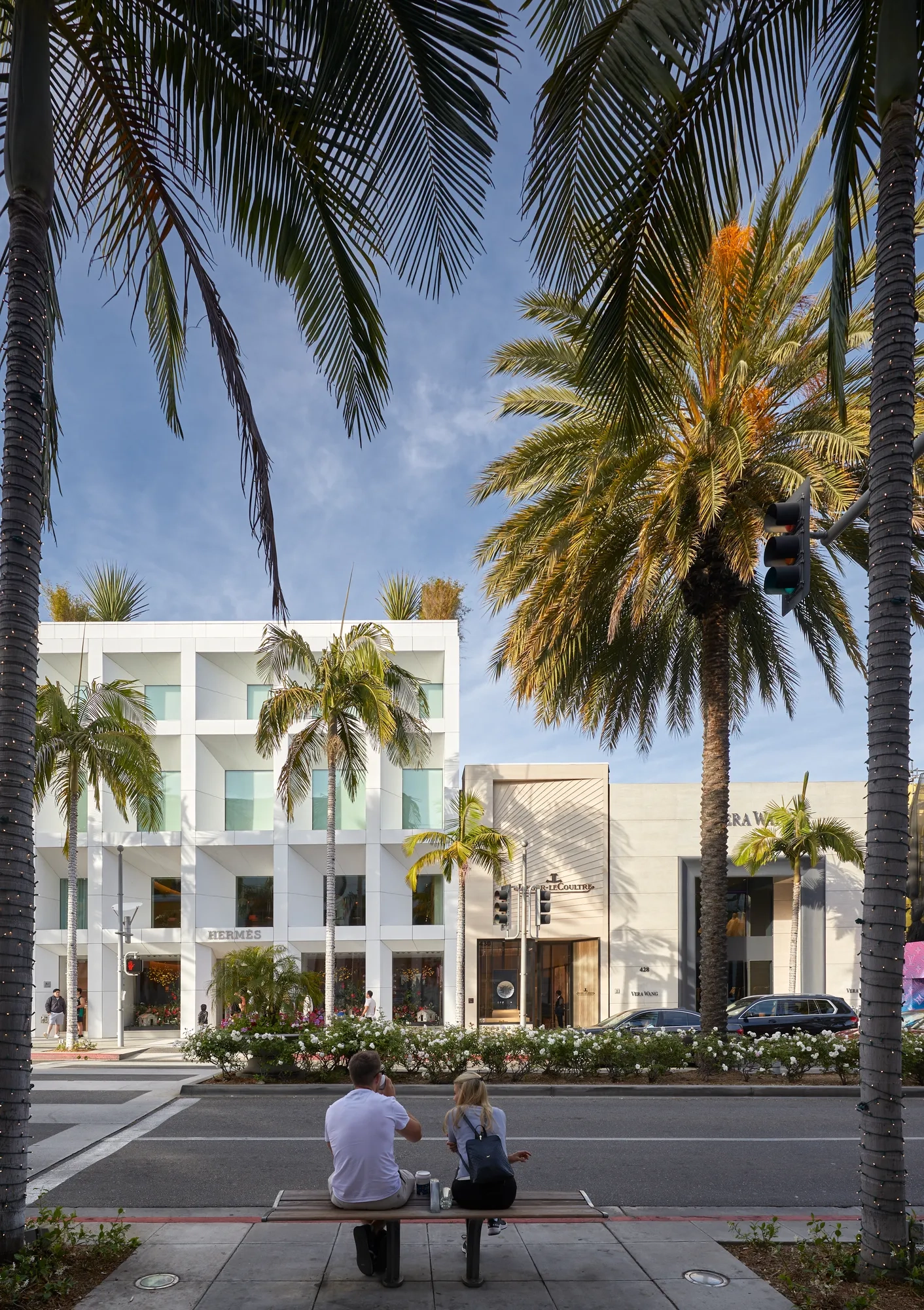 Two people on a bench facing Hermès and Jaeger-LeCoultre stores across Rodeo Drive, framed by palm fronds.