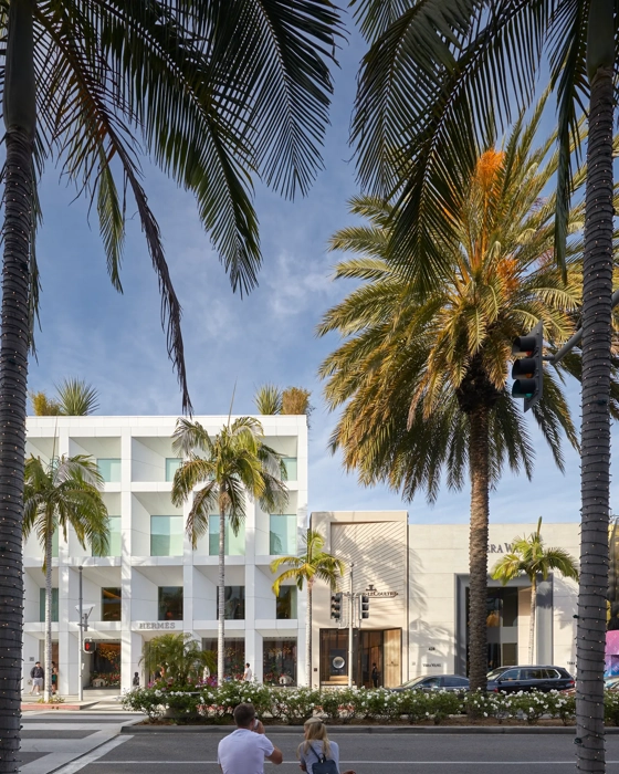 Two people on a bench facing Hermès and Jaeger-LeCoultre stores across Rodeo Drive, framed by palm fronds.