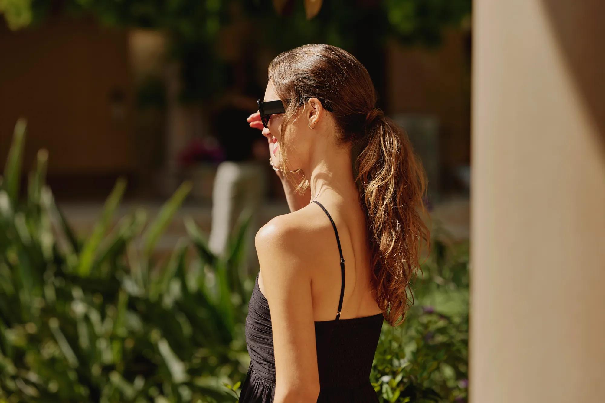Person in a black dress and sunglasses walks through a sunlit garden terrace surrounded by lush greenery and columns.