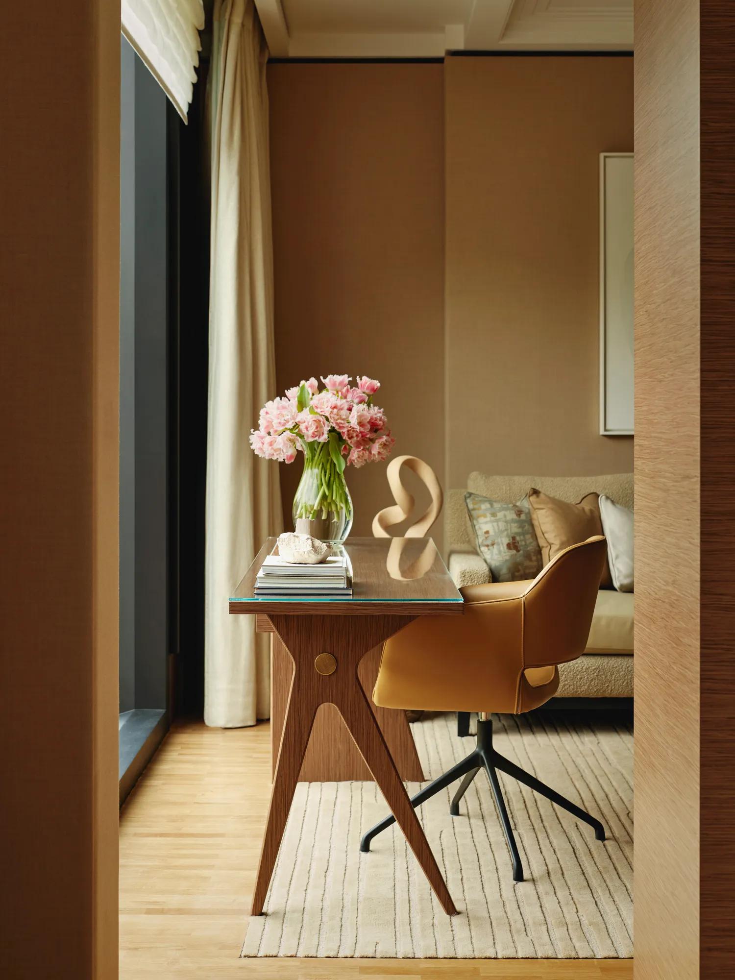 Writing desk with a leather swivel chair and vase of pink flowers, set by a window with a sofa in the background.