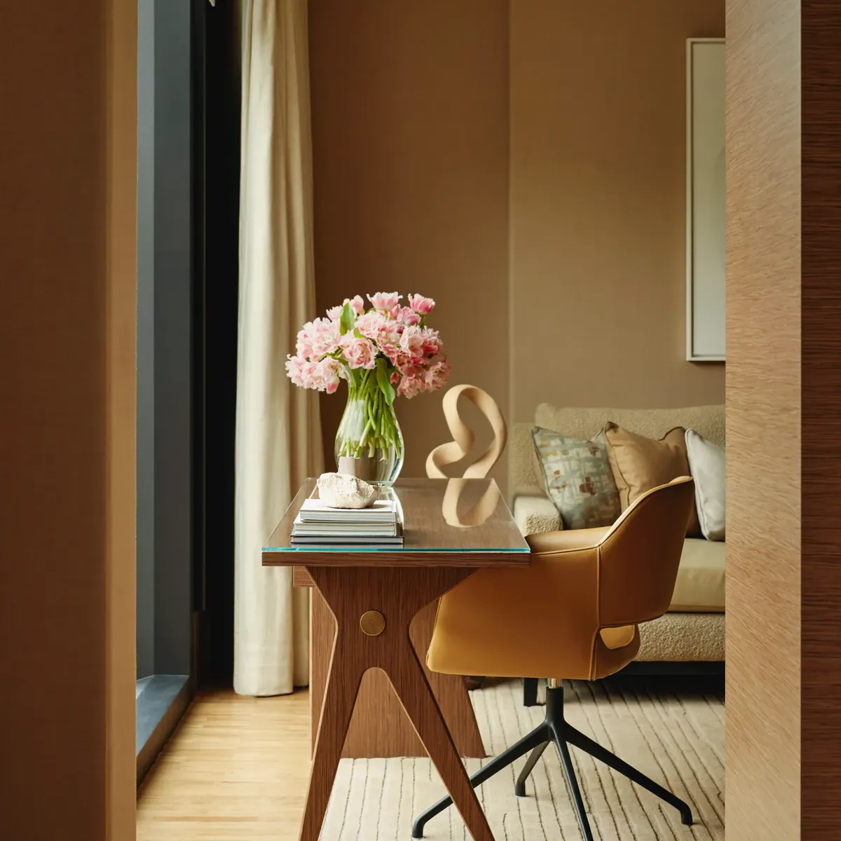 Writing desk with a leather swivel chair and vase of pink flowers, set by a window with a sofa in the background.