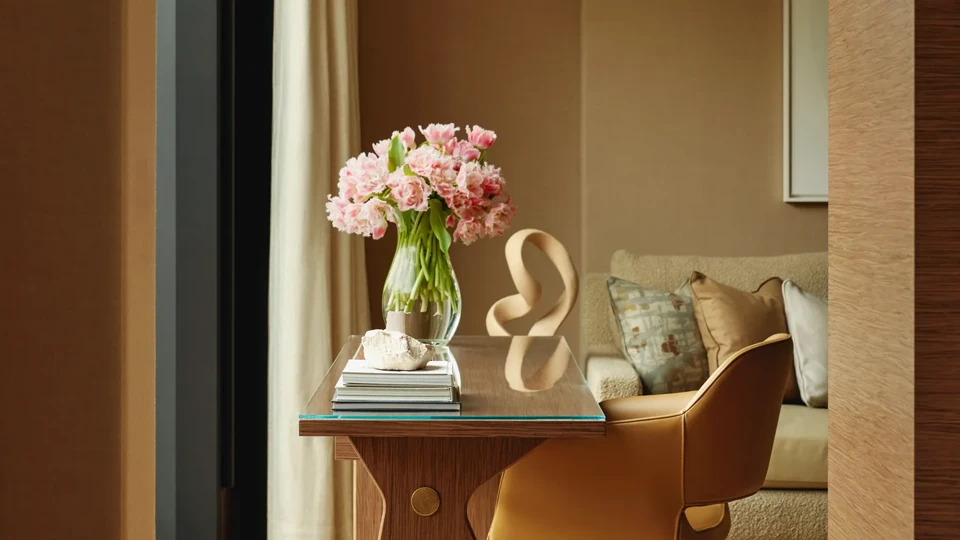 Writing desk with a leather swivel chair and vase of pink flowers, set by a window with a sofa in the background.