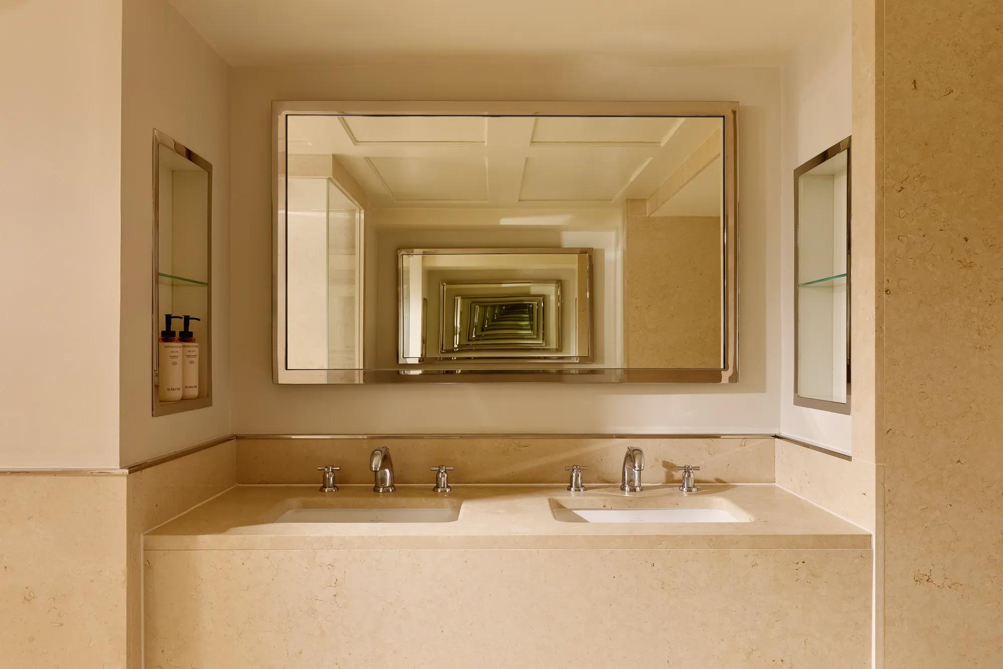 Cream marble bathroom with a double vanity and two sinks, chrome fixtures, and a large mirror creating a repeating reflection effect across the space.