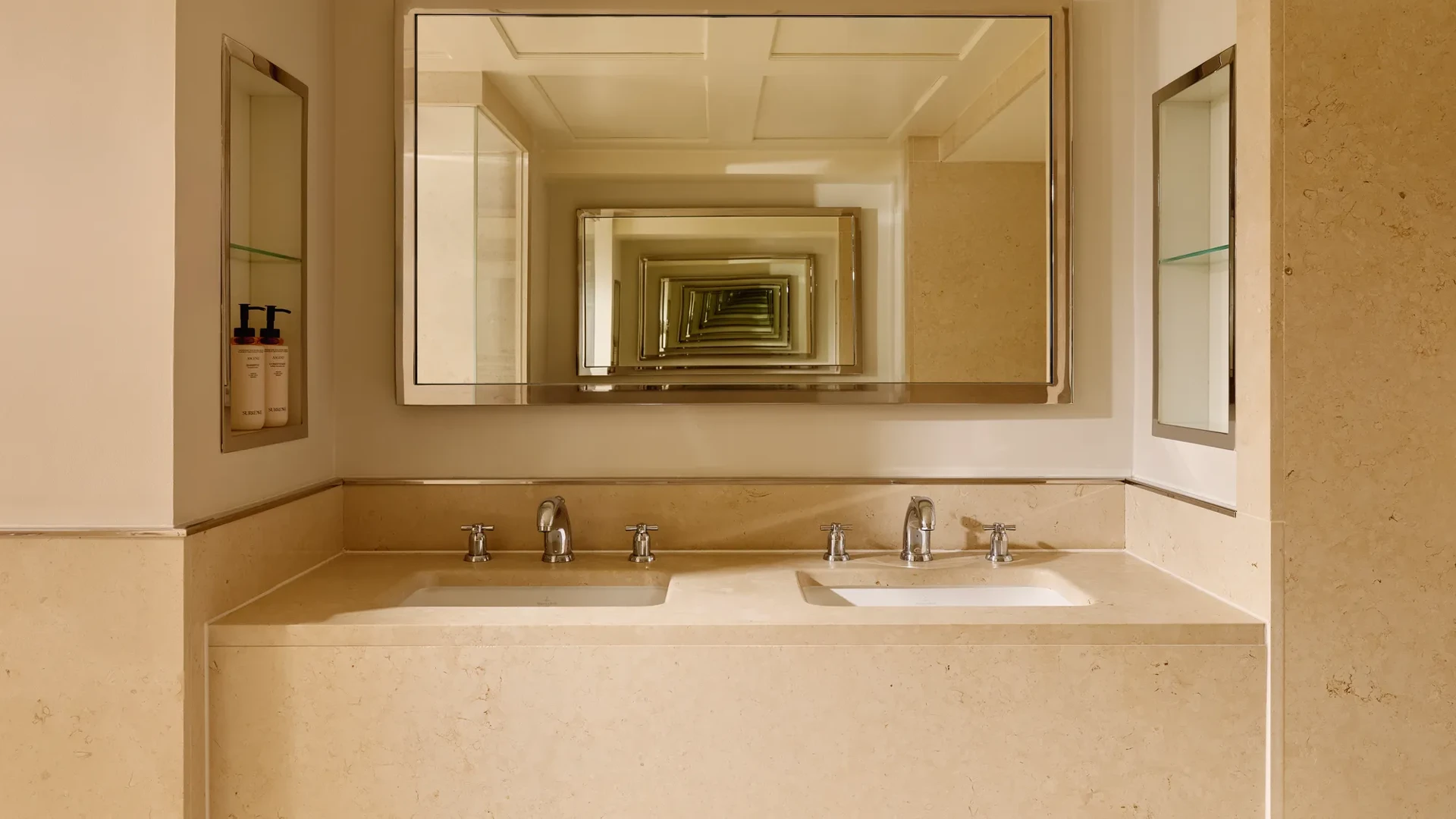 Cream marble bathroom with a double vanity and two sinks, chrome fixtures, and a large mirror creating a repeating reflection effect across the space.