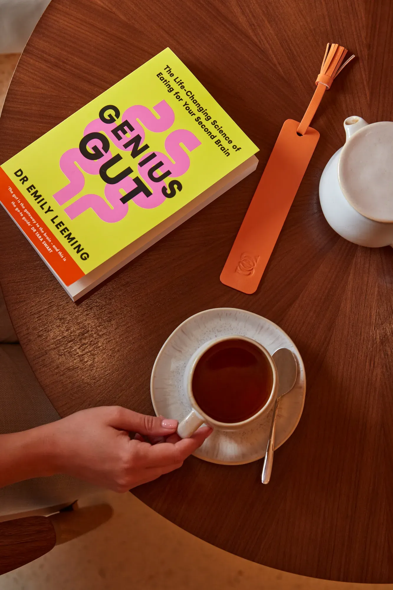 Top-down view of a hand holding a coffee cup beside the book Genius Gut on a round wooden table.