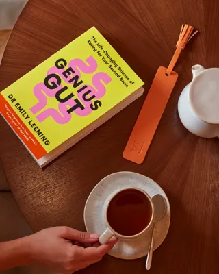 Top-down view of a hand holding a coffee cup beside the book Genius Gut on a round wooden table.