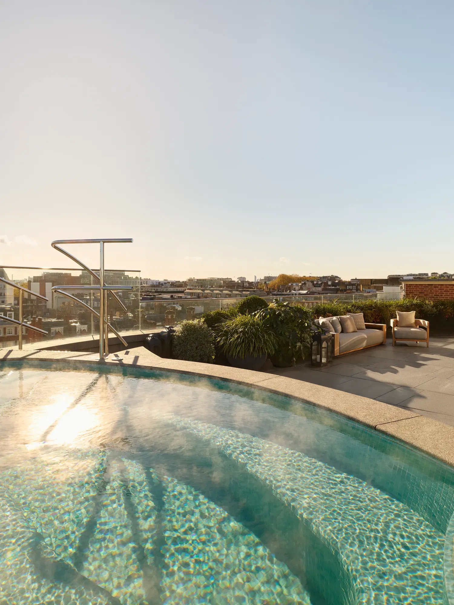 Rooftop terrace with a curved heated pool surrounded by greenery, featuring cushioned outdoor seating and panoramic city views under a clear blue sky.