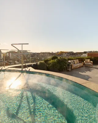 Rooftop terrace with a curved heated pool surrounded by greenery, featuring cushioned outdoor seating and panoramic city views under a clear blue sky.