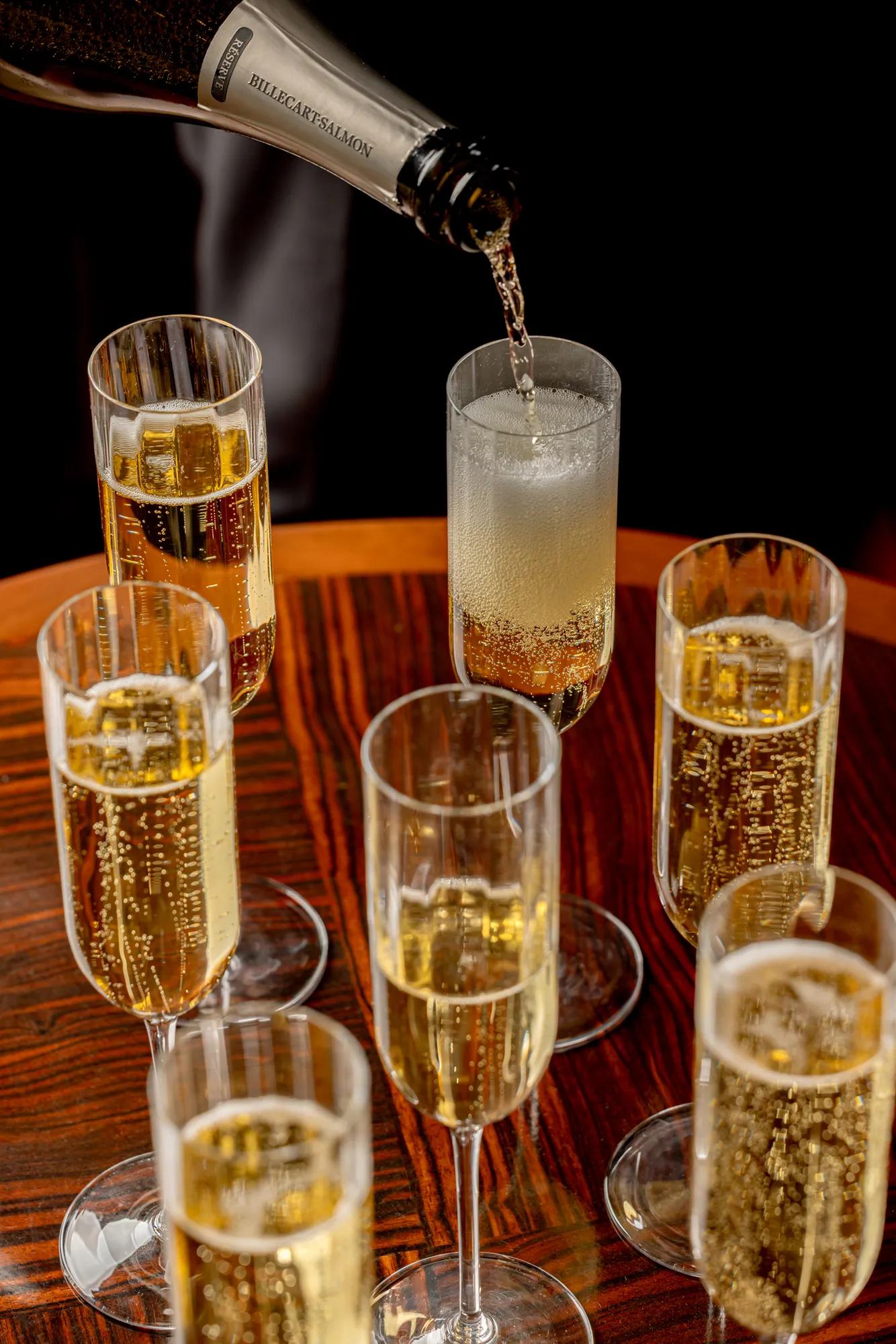 Champagne being poured into a tall flute, surrounded by filled glasses on a polished wooden table.