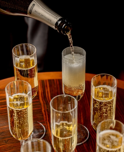 Champagne being poured into a tall flute, surrounded by filled glasses on a polished wooden table.