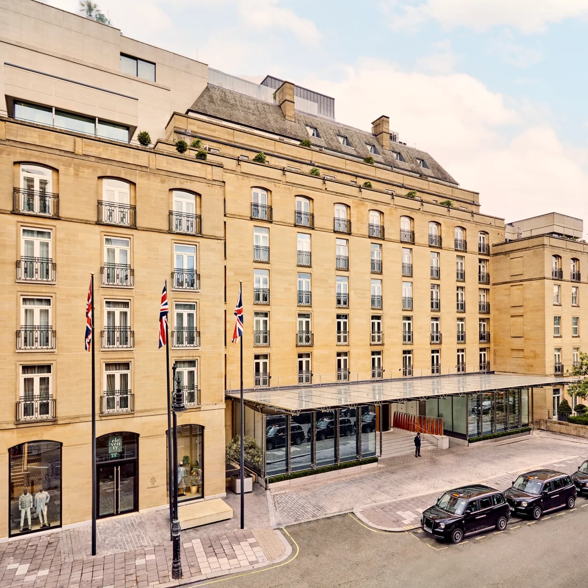Elegant sandstone hotel façade with Union Jack flags and glass entrance canopy on a quiet London street with black cabs parked outside.