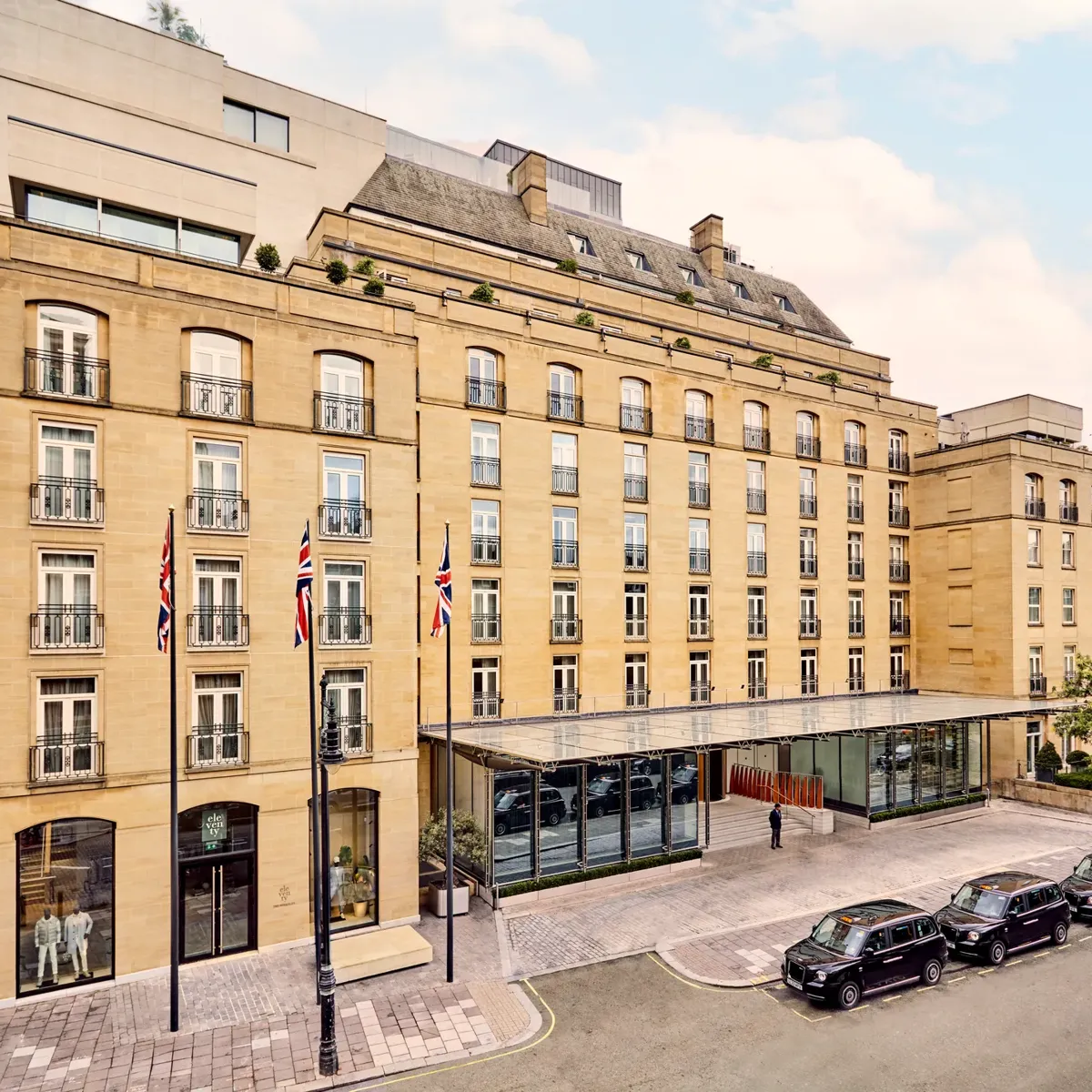 Elegant sandstone hotel façade with Union Jack flags and glass entrance canopy on a quiet London street with black cabs parked outside.