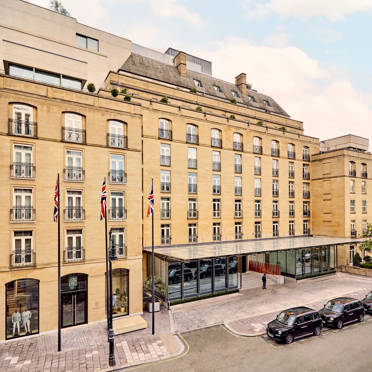 Elegant sandstone hotel façade with Union Jack flags and glass entrance canopy on a quiet London street with black cabs parked outside.