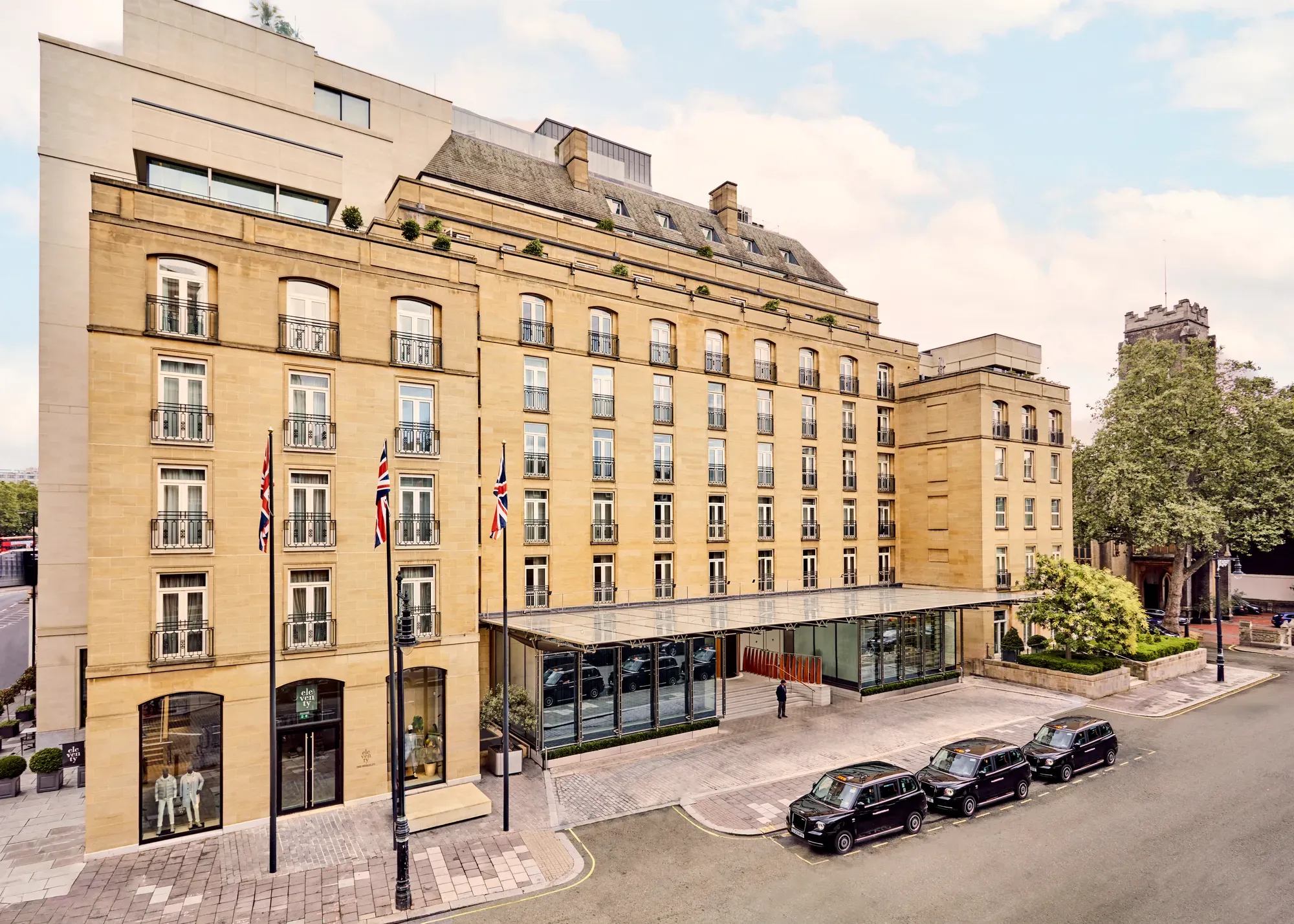 Elegant sandstone hotel façade with Union Jack flags and glass entrance canopy on a quiet London street with black cabs parked outside.