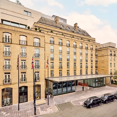 Elegant sandstone hotel façade with Union Jack flags and glass entrance canopy on a quiet London street with black cabs parked outside.