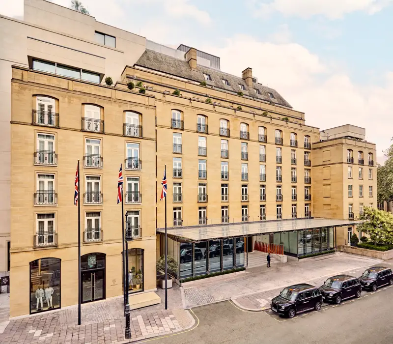 Elegant sandstone hotel façade with Union Jack flags and glass entrance canopy on a quiet London street with black cabs parked outside.