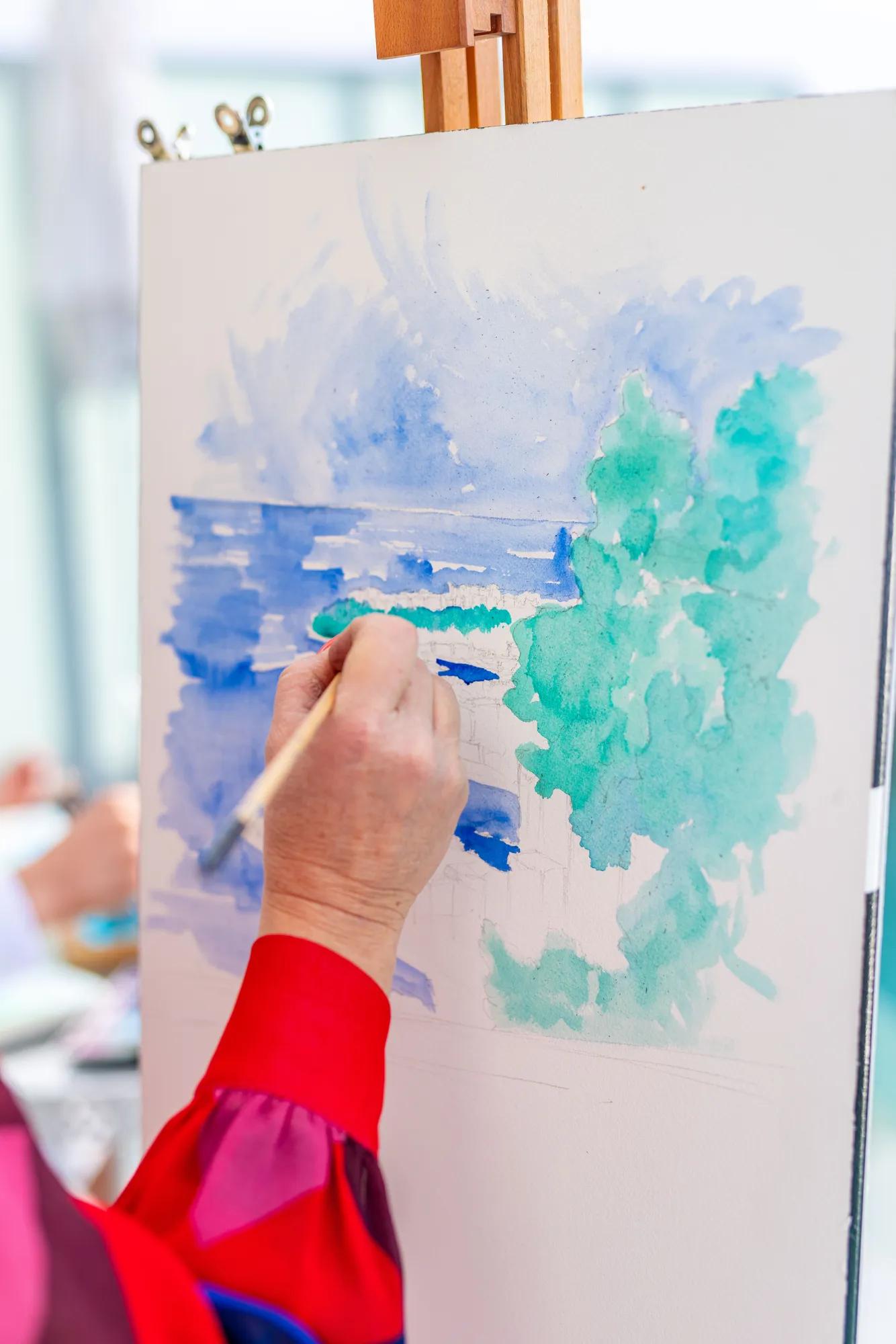 Gros plan d’une main peignant une aquarelle représentant un paysage marin avec des arbres en tons verts et bleus sur une toile, à l’aide d’un pinceau fin.