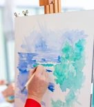 Gros plan d’une main peignant une aquarelle représentant un paysage marin avec des arbres en tons verts et bleus sur une toile, à l’aide d’un pinceau fin.