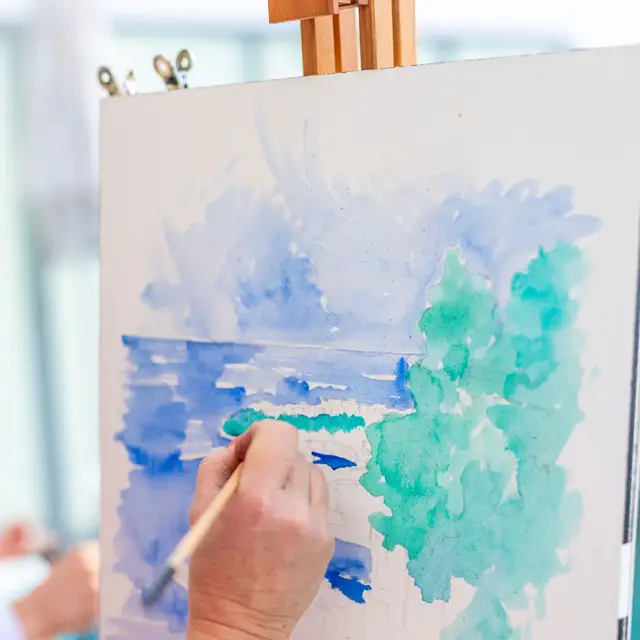 Gros plan d’une main peignant une aquarelle représentant un paysage marin avec des arbres en tons verts et bleus sur une toile, à l’aide d’un pinceau fin.