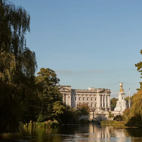 View across St James’s Park lake towards Buckingham Palace and Victoria Memorial, framed by trees in warm evening light.