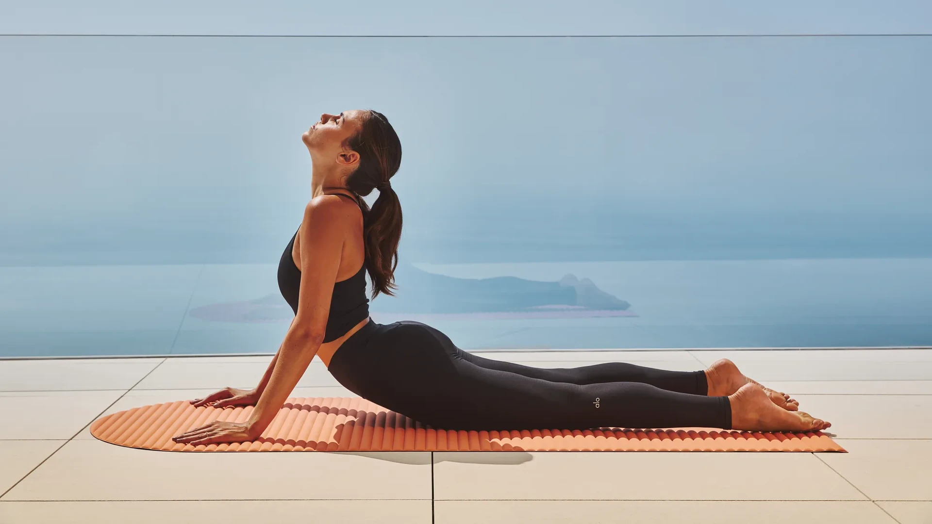 Person practising upward-facing dog yoga pose on a mat, overlooking the sea through a glass balustrade.