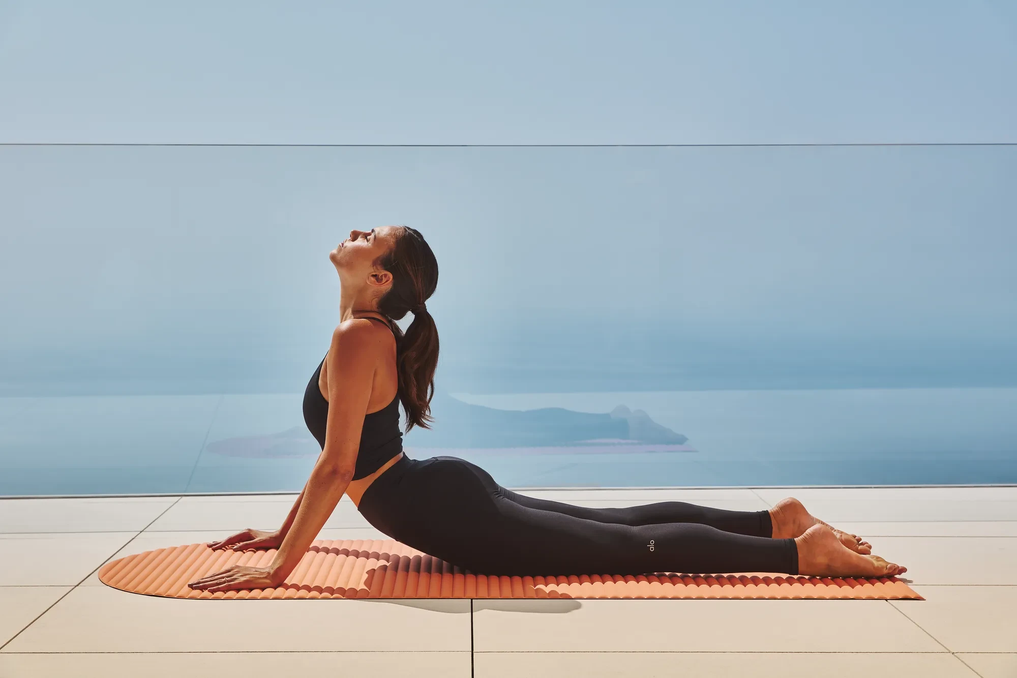 Person practising upward-facing dog yoga pose on a mat, overlooking the sea through a glass balustrade.
